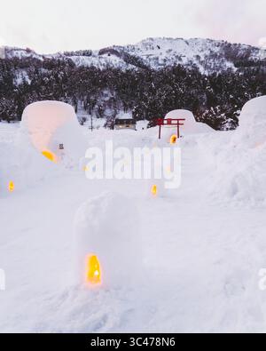 La vista delle cupole di neve scintillanti e di un cancello torii rosso si staglia contro la neve bianca e gli alberi scuri nel villaggio innevato di Kamakura, Nagano, Giappone. Foto Stock