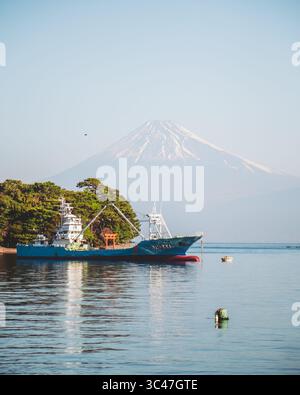 Heda, Giappone - 1 marzo 2025: Vista di un peschereccio che riposa pacificamente nelle tranquille acque della baia, con il maestoso Monte Fuji che si innalza in una distanza nebbiosa. Foto Stock