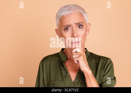 Affascinante donna matura con i capelli bianchi corti che indossa una camicia verde cachi, in posa su uno sfondo beige Foto Stock