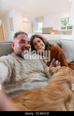 Coppia che indossa maglioni grossi seduti sul divano in una cucina aperta con lo smartphone che coccola il cane Foto Stock