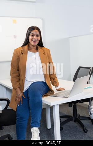 Donna afroamericana di medie dimensioni seduta sul bordo della scrivania nella sala meeting con computer portatile aperto e lavagna bianca Foto Stock
