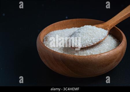 Zucchero granulato bianco in una ciotola di legno con un cucchiaio di legno pieno di zucchero, appoggiato su uno sfondo nero. Ideale per confezionamento di alimenti, cucina V Foto Stock