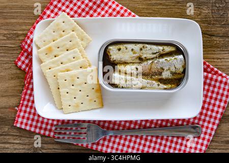 Una scatola aperta di sardine in olio, servita accanto a una pila di cracker quadrati su un piatto bianco rettangolare, uno spuntino semplice e nutriente. Foto Stock