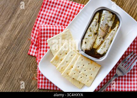 Vista dall'alto di una stagnola aperta di sardine in olio, servita accanto a una pila di cracker quadrati su un piatto bianco rettangolare. Foto Stock