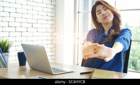 Donna che stringe il braccio sollevato, seduta in posizione non corretta, scrivania da ufficio domestico. Lato posteriore di giovane donna asiatica stanca da lavoro corpo stress mal di schiena ufficio sy Foto Stock