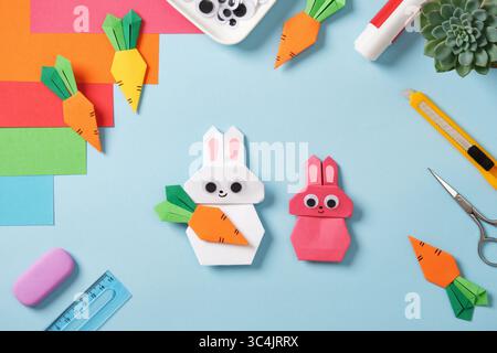 Vista dall'alto del desktop creativo. Coniglietti origami rosa e bianco con carote arancioni. Forniture artistiche fai da te: Carta, forbici, coltello per cartoleria, pennarelli Foto Stock