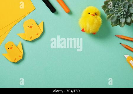 Inquadra su sfondo turchese con due pulcini origami di carta gialla, pulcino giocattolo giallo soffice. Forniture d'arte: Matite, pennarelli, coltello da disegno. PL Foto Stock