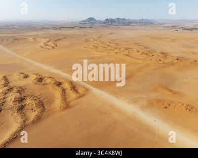 Veduta aerea di un paesaggio arido e soleggiato dove la strada pallida taglia le dune ocra verso montagne lontane, deserto del Namib, Namibia. Foto Stock