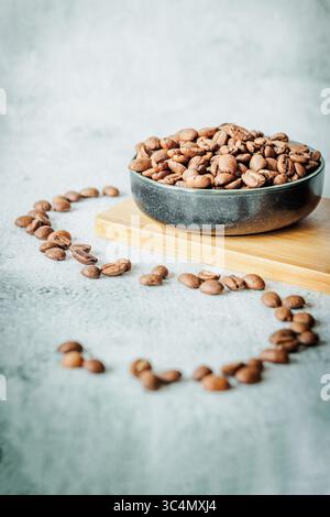 Chicchi di caffè tostati in una ciotola di ceramica scura posta su una tavola di bambù, incorniciata da una dispersione circolare di chicchi su cemento chiaro, che trasporta un caldo caffè artigianale Foto Stock
