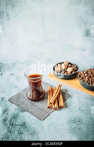 Bicchiere ricurvo di caffè nero con bastoncini di cannella, ciotole di chicchi tostati e spezie riscaldanti su tavola di bambù, modellato su pietra testurizzata con grande ariosità Foto Stock