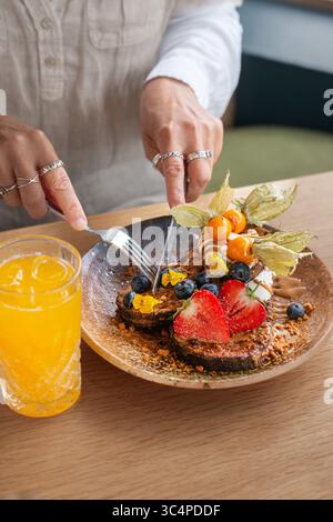 Primo piano di una persona che taglia una fetta di pane tostato ricoperto di frutti di bosco freschi e panna utilizzando una forchetta e un coltello. Colazione salutare o brunch fatto in casa Foto Stock