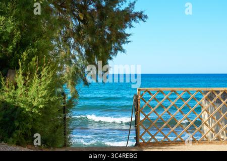 Splendida vista del mare blu e delle onde attraverso un cancello giardino in legno. Paesaggio costiero mediterraneo panoramico in un giorno d'estate soleggiato Foto Stock