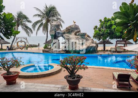 Questo invitante resort sulla spiaggia dispone di una piscina cristallina, cascata di roccia naturale, palme e serene vedute dell'oceano con accoglienti punti lounge Foto Stock