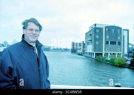 Ritratto di un uomo, 00-00-1999 Whizgle News, Dutch Desk, Paesi Bassi, 1950-2000 Una persona sorridente si trova vicino a un corpo d'acqua, con edifici industriali sullo sfondo, incorniciati da un cielo nuvoloso. Foto Stock