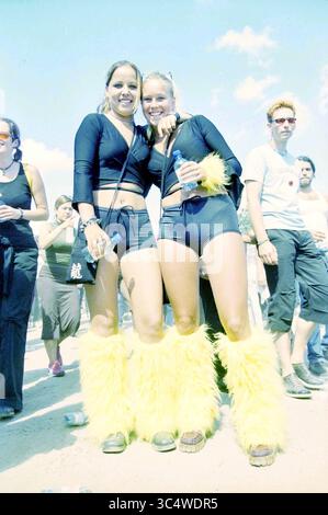 Amanti della Dance Valley, 03-08-2001 Whizgle News, Dutch Desk, Paesi Bassi, 1950-2000 due donne stanno insieme, sorridono con fiducia indossando abiti coordinati che presentano giocosi e soffici scaldasci gialli. L'atmosfera vivace include altri frequentatori di festival sullo sfondo, creando un senso di gioia e cameratismo. Foto Stock