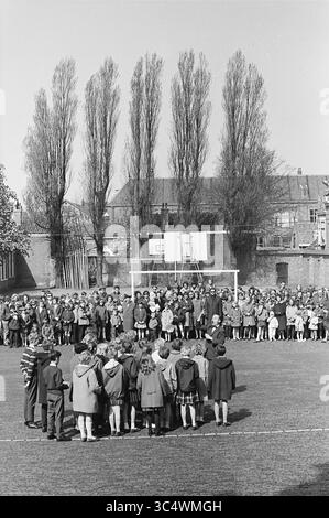 Coro per bambini con pubblico all'esterno, 05-05-1964 Whizgle News, Dutch Desk, Paesi Bassi, 1950-2000 Un grande gruppo di scolari si riunisce in un parco giochi, con alcuni in piedi in cerchio mentre altri guardano dai lati. Alberi alti fiancheggiano lo sfondo e un canestro da basket si erge in modo prominente. La scena cattura un vivace evento scolastico all'aperto, pieno di energia giovanile e cameratismo. Foto Stock