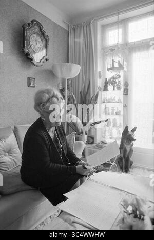 Donna con cane in soggiorno, Haarlem, Paesi Bassi, 04-05-1981 Whizgle News, scrivania olandese, i Paesi Bassi, 1950-2000 una donna anziana sedeva comodamente su un divano, leggendo documenti, con un curioso gatto nelle vicinanze, il tutto circondato da un accogliente soggiorno decorato con oggetti decorativi e un orologio da parete. Foto Stock