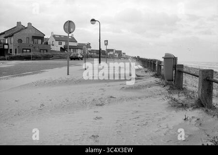 Sabbia su Boulevard Zandvoort., Zandvoort, Zandvoort, 04-01-1987 Whizgle News, Dutch Desk, i Paesi Bassi, 1950-2000 Una strada costiera deserta costeggiata da case, con una panchina, un cartello stradale e una cassetta postale, è parzialmente coperta di sabbia, suggerendo una recente tempesta o un'alta marea. La scena trasmette un senso di tranquillità e abbandono, con colline lontane e un cielo nuvoloso in alto. Foto Stock