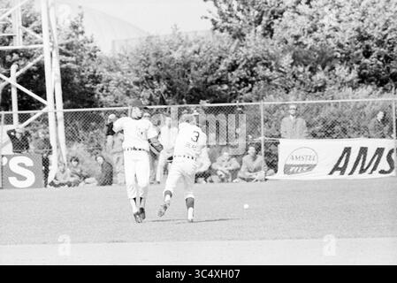 Baseball Paesi Bassi - Detroit ITM, 27-06-1971 Whizgle News, Dutch Desk, Paesi Bassi, 1950-2000 due giocatori di baseball sembrano saltare contemporaneamente nel tentativo di prendere una palla durante una partita, mentre gli spettatori guardano da dietro una recinzione. La scena cattura un momento di azione ed eccitazione sul campo. Foto Stock