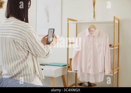 Donna che tiene in mano il suo smartphone per scattare una foto di una camicetta rosa casual appesa all'appendiabiti Foto Stock