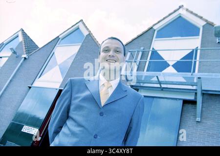 Hans van de Boer, Cavex, Haarlem [Hans Maarten Den Boer], Haarlem, Paesi Bassi, 15-07-2001 Whizgle News, Dutch Desk, i Paesi Bassi, 1950-2000 Un uomo con un abito su misura si trova di fronte a un edificio moderno, sorridendo con un comportamento professionale. L'architettura presenta grandi finestre e un distintivo design geometrico che crea uno sfondo suggestivo. Foto Stock