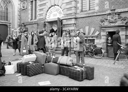 Gruppo di giovani con bagagli da viaggio, di fronte a Vleeshal Haarlem (in occasione del festival Hartewens), Haarlem, Nederland, 00-00-1966 Whizgle News, Dutch Desk, i Paesi Bassi, 1950-2000 Una vivace scena fuori da un edificio storico, dove un gruppo di persone viene riunito con varie valigie e borse. Alcuni individui sono visti entrare e uscire dall'edificio, mentre altri si impegnano nella conversazione, creando un'atmosfera vivace. Le biciclette sono parcheggiate nelle vicinanze, aggiungendo fascino all'ambiente. Foto Stock
