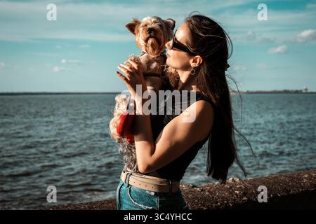 Una donna e lo Yorkshire terrier condividono un dolce momento in acqua in una giornata di sole. Una ragazza abbraccia un piccolo cane vicino all'acqua, godendosi un momento di affetto. Foto Stock