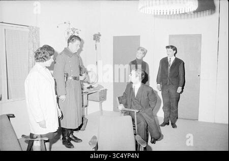 Spettacolo teatrale Whizgle News, Dutch Desk, Paesi Bassi, 1950-2000 Un gruppo di cinque persone si impegna in una seria discussione all'interno di una stanza scarsamente arredata, evidenziando una tensione tra una figura in uniforme e gli altri. L'impostazione suggerisce un contesto formale o ufficiale, con un elemento di autorità presente. Foto Stock