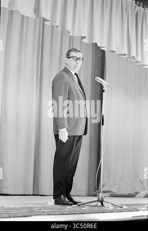 Ria Valk con chitarra Andre Meurs, Cabaret, 13-04-1966 Whizgle News, Dutch Desk, Paesi Bassi, 1950-2000 Un uomo si trova davanti a un microfono sul palco, vestito con un abito con occhiali, rivolgendosi a un pubblico da dietro uno stand. Lo sfondo presenta tende drappeggiate, creando un'atmosfera formale per il suo discorso. Foto Stock