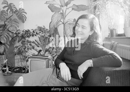 Ritratto di una donna Whizgle News, Dutch Desk, Paesi Bassi, 1950-2000 Una giovane donna irradia di gioia, seduta comodamente in un accogliente spazio abitativo adornato da piante lussureggianti. Indossa un elegante maglione scuro e una gonna al ginocchio, trasudando un fascino rilassato mentre si diverte a trascorrere un momento di svago accanto a un televisore d'epoca. Foto Stock