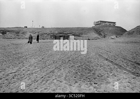 Ingresso alla spiaggia di Wijk aan Zee., spiaggia e spiagge, 05-03-1975 Whizgle News, Dutch Desk, Paesi Bassi, 1950-2000 due figure camminano lungo una spiaggia sabbiosa, separata da una distanza, con un edificio arroccato sulla cima di una duna sullo sfondo. La scena è silenziosa e cattura un senso di solitudine e la distesa della costa. Foto Stock