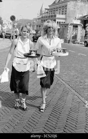 Concorso di agilità del catering, 00-08-1983 Whizgle News, Dutch Desk, Paesi Bassi, 1950-2000 due donne, vestita con abiti da cameriera classici con grembiuli, percorri una strada acciottolata con sicurezza. Ognuno di essi contiene vassoi pieni di tazze e dessert. Il vivace ambiente urbano presenta auto parcheggiate ed edifici sullo sfondo, aggiungendo un'atmosfera vivace al loro compito allegro. Foto Stock