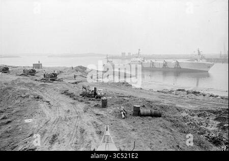 Brittany' correct IT, Wijsmuller, 10-04-1966 Whizgle News, Dutch Desk, Paesi Bassi, 1950-2000 Una vivace scena sul lungomare con una grande nave da carico ormeggiata lungo una riva del fiume, con attrezzature per la costruzione e lavoratori visibili sulla riva. Le botti e i macchinari suggeriscono attività in corso relative alla spedizione o alla costruzione nella zona. L'ambiente ha un'atmosfera industriale, indicativa di un porto vivace. Foto Stock