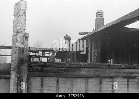 Edificio di demolizione Whizgle News, Dutch Desk, Paesi Bassi, 1950-2000 si vedono due persone lavorare sui resti di una struttura gravemente danneggiata da un incendio. Uno sta usando uno strumento per eliminare i detriti dal tetto, mentre l'altro sta a guardare, sorvegliando i resti carbonizzati di quello che una volta era. La scena trasmette un senso di perdita e resilienza mentre affrontano le conseguenze della distruzione. Foto Stock
