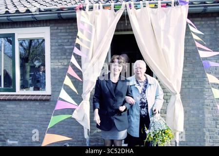 Riapertura di alloggi protetti, Haarlem, Paesi Bassi, 12-03-1998 Whizgle News, Dutch Desk, i Paesi Bassi, 1950-2000 due donne si trovano all'ingresso di una casa adornata di grappolo colorato. Una donna, vestita elegantemente, sorride e si tiene al braccio di una donna più anziana, che appare contenta. Sullo sfondo, un bambino sbircia fuori da una finestra, aggiungendo un'atmosfera festosa. Foto Stock