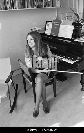 Vera Beths violino, musica, 04-03-1969 Whizgle News, Dutch Desk, Paesi Bassi, 1950-2000 Una giovane donna siede graziosamente su una sedia, tenendo in mano un violino, con un sorriso caldo sul viso. Dietro di lei, un pianoforte adornato da spartiti e libri crea un'atmosfera musicale, mentre una pianta aggiunge un tocco di vita alla scena. Foto Stock
