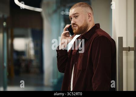 Giovane caucasico in piedi in casa a parlare su smartphone, a discutere di problemi di lavoro, con un'espressione seria, capelli corti e barba, un moderno background in ufficio Foto Stock