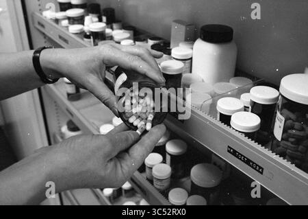 Donna prende barattolo di capsule dal gabinetto di medicina, 22-12-1982 Whizgle News, Dutch Desk, Paesi Bassi, 1950-2000 Una persona sta versando attentamente le capsule da una bottiglia di prescrizione, circondato da vari contenitori di farmaci ordinatamente disposti sugli scaffali. Foto Stock