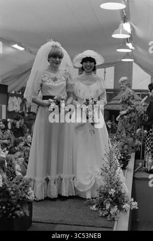 Sfilata di moda Liba Spring Fair Haarlem, sfilata di moda, fiera di primavera, 08-03-1976 Whizgle News, Dutch Desk, i Paesi Bassi, 1950-2000 due spose camminano lungo una passerella, sorridendo mentre espongono i loro eleganti abiti da sposa. Uno indossa un abito classico con un velo, mentre l'altro sfoggia un abito d'ispirazione vintage decorato con un grande cappello. Sullo sfondo, il pubblico guarda attentamente, mettendo in evidenza l'atmosfera festosa dell'evento. Foto Stock
