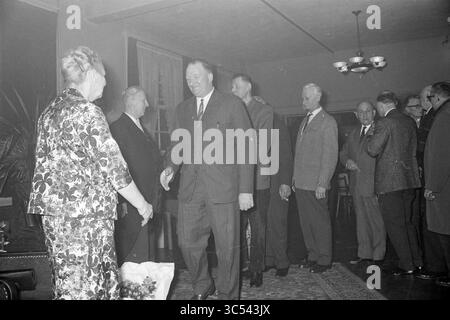 Omaggio a Lord Captain a Groenendaal, omaggio, 31-03-1964 Whizgle News, Dutch Desk, Paesi Bassi, 1950-2000 Un gruppo di uomini in abbigliamento formale sono riuniti in una stanza poco illuminata, impegnati in una conversazione. Un uomo estende la mano verso una donna vestita con un abito floreale, mentre altri sembrano osservare l'interazione. L'atmosfera suggerisce un evento formale o una riunione. Foto Stock