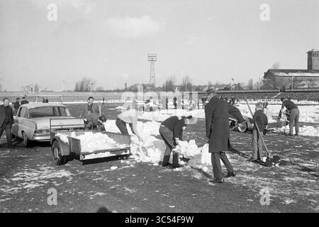 Clearing Snow in Haarlem Grounds, Football Haarlem, Work, 04-02-1968 Whizgle News, Dutch Desk, i Paesi Bassi, 1950-2000 Un gruppo di persone vestite di abbigliamento invernale sta sgombrando una zona innevata, utilizzando pale e rimorchi per trasportare la neve. Sullo sfondo, sono visibili strutture e una torre, mentre i veicoli parcheggiati sono visti nelle vicinanze, indicando uno sforzo organizzato per rimuovere la neve dal suolo. Foto Stock