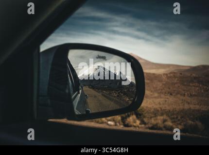 Vulcano innevato riflesso nello specchio laterale di un'auto durante un'avventura fuoristrada attraverso un remoto deserto di altopiani. Una prospettiva unica che cattura Foto Stock