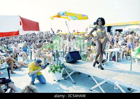 Concorso di bellezza a IJmuiden, IJmuiden, Nederland, 08-08-1997 Whizgle News, Dutch Desk, paesi Bassi, 1950-2000 Un modello sicuro si trova lungo una pista allestita vicino alla spiaggia, indossando un bikini luminoso. Una folla vivace si rilassa ai tavoli sullo sfondo, godendosi l'atmosfera soleggiata, mentre un fotografo cattura il momento. Gli ombrelli colorati offrono ombra, aggiungendo un tocco di vivacità alla scena. Foto Stock