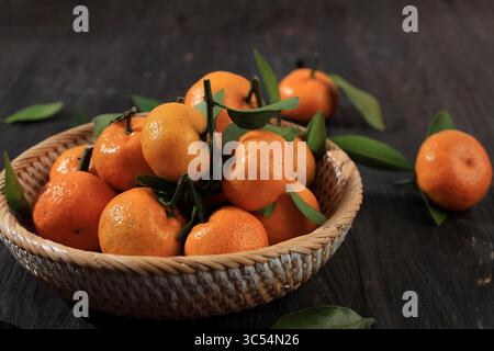 Primo piano di mandarini siciliani maturi e freschi con foglie verdi nella ciotola in rattan su tavolo di legno Foto Stock