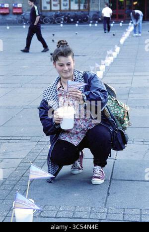 Campagna pubblicitaria dell'operatore ferroviario Amsterdam - IJmuiden Lovers Rail con un nastro di secchi di sabbia pieni con la bandiera "IJmuiden non scoperto" da Dam alla stazione centrale, Amsterdam, Paesi Bassi, 30-07-1997 Whizgle News, Dutch Desk, i Paesi Bassi, 1950-2000 Anni Un giovane, vestito con un abbigliamento casual, accovacciato su un marciapiede, tenendo in mano un container e posizionando una piccola bandiera di fronte a loro. Sullo sfondo, le persone camminano, e una linea di bandiere è visibile, creando un senso di attività comunitaria. Foto Stock
