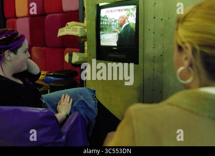 Attacco a PIM Fortuyn studio Haarlem, Haarlem, Paesi Bassi, 05-05-2002 Whizgle News, Dutch Desk, i Paesi Bassi, 1950-2000 Una giovane donna siede comodamente su una sedia colorata, guardando un programma su una piccola televisione, mentre un'altra donna, leggermente allontanata, osserva lo schermo. L'ambiente presenta uno sfondo vibrante e posti a sedere informali, creando un'atmosfera rilassata. Foto Stock