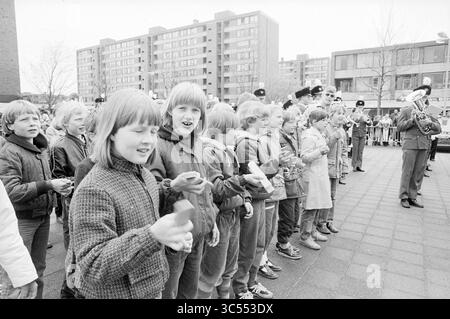 Klepper march presso l'ufficio cittadino B'wijk, Queen's Day, Beverwijk, Paesi Bassi, 30-04-1982 Whizgle News, Dutch Desk, Paesi Bassi, 1950-2000 un gruppo di bambini partecipa con entusiasmo a un vivace evento all'aperto, tenendo piccoli oggetti mentre assistono a un'esibizione di una banda in marcia. Le loro espressioni mostrano eccitazione e curiosità, mentre una folla di spettatori si riunisce sullo sfondo vicino a edifici moderni. Foto Stock