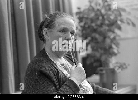 Sig.ra De Haan, ex governante Menten, Persons, 26-11-1976 Whizgle News, Dutch Desk, i Paesi Bassi, 1950-2000 Una donna contemplativa è seduta, tenendo leggermente un paio di occhiali in bocca, apparentemente persa nel pensiero. Ha un'espressione delicata ed è vestita con una camicetta a motivi geometrici sotto un accogliente cardigan, con tende e una pianta in vaso che incornicia delicatamente lo sfondo. Foto Stock