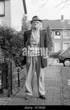Ritratto dell'uomo con cappello Whizgle News, Dutch Desk, Paesi Bassi, 1950-2000 un uomo anziano si erge su un sentiero di pietra, vestito da un blazer elegante con un maglione a motivi geometrici sotto. Indossa una fedora e ha una sigaretta sporca dalle labbra. Dietro di lui, le case residenziali e il verde creano uno sfondo suburbano, aggiungendo un tocco di carattere alla scena. Foto Stock
