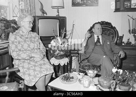 60 anni di coppia nuziale Koningshoeve e Spronk, matrimonio, 27-12-1976 Whizgle News, Dutch Desk, Paesi Bassi, 1950-2000 un uomo e una donna siedono comodamente in un accogliente soggiorno, circondato da piante e arredi vintage. La donna, vestita con un abito a fantasia, sorride mentre si impegna con l'uomo, che è seduto su una sedia ornata, tenendo un sigaro. Un piccolo tavolino da caffè di fronte a loro è decorato con spuntini e oggetti decorativi, mentre un televisore aggiunge all'atmosfera nostalgica dello spazio. Foto Stock